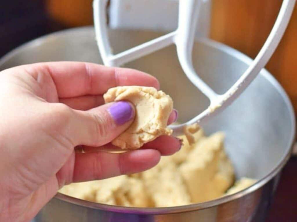 A hand with purple nail polish holds a piece of cookie dough in front of a stand mixer with a paddle attachment and more dough inside the bowl, while the aroma of crockpot Mexican beef lingers warmly in the background.