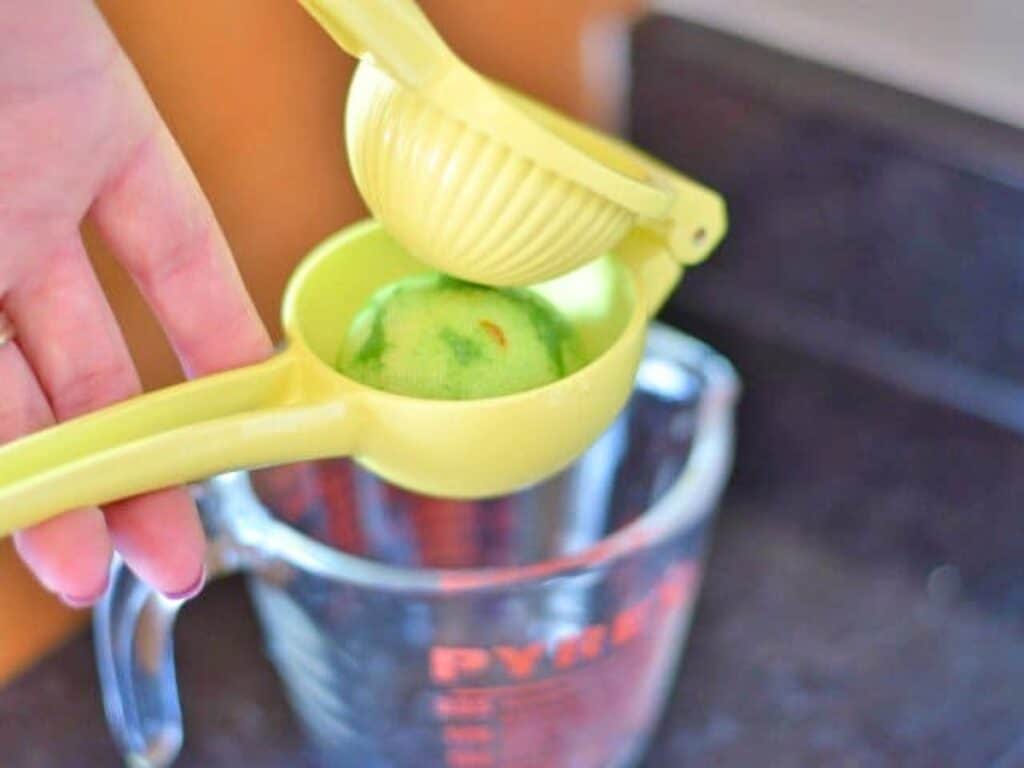 A hand squeezes a green citrus fruit with a yellow handheld juicer over a glass measuring cup on a kitchen counter, preparing fresh juice for crockpot Mexican beef.