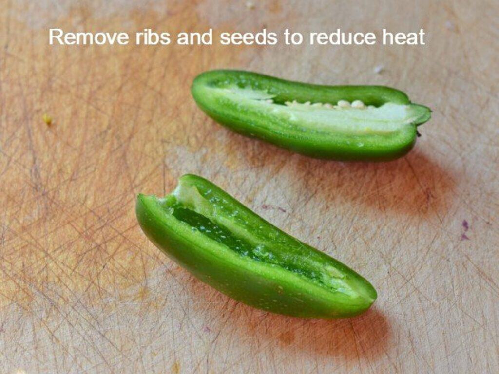 Two halves of a green chili pepper with visible seeds rest on a wooden cutting board—perfect for adding just the right kick to your crockpot Mexican beef. Text reads, “Remove ribs and seeds to reduce heat.”.