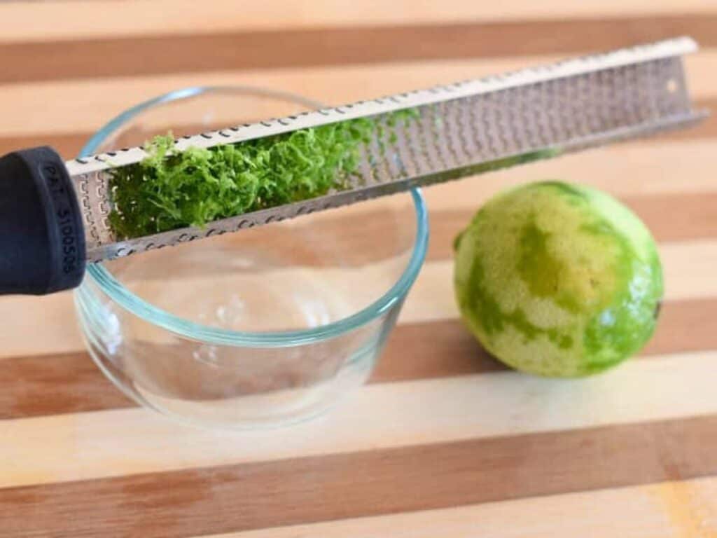 A microplane grater is being used to zest a lime over a small glass bowl on a striped wooden surface, preparing fresh ingredients for crockpot Mexican beef.