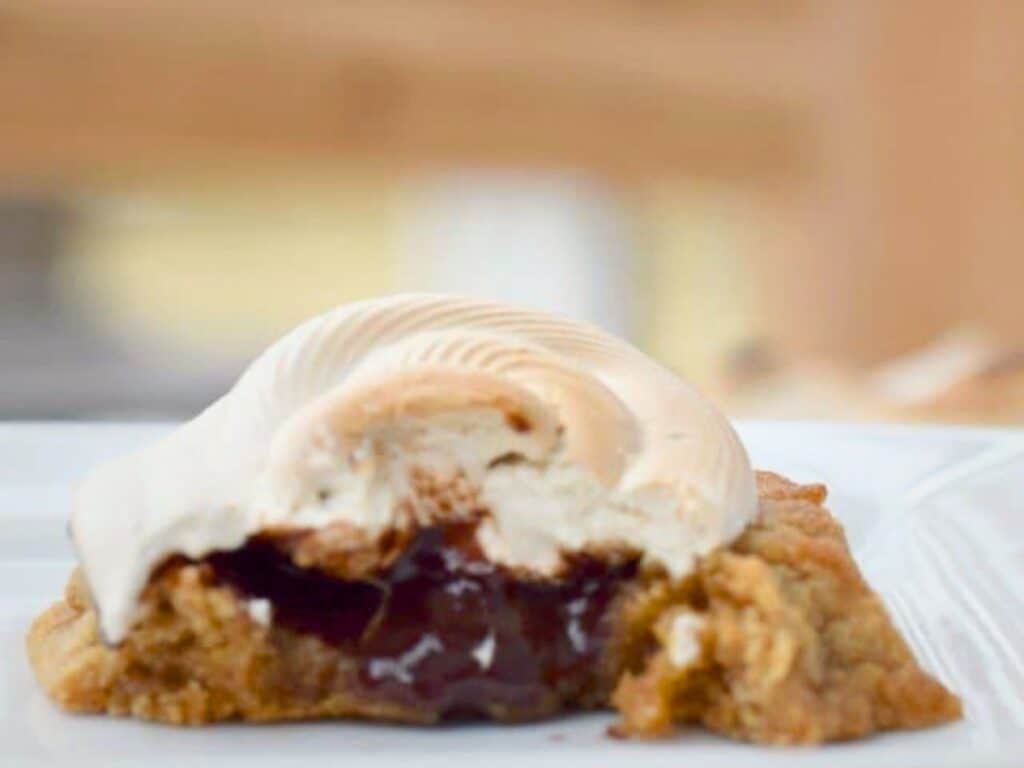 A close-up of a s&rsquo;mores cookie with a golden meringue topping and a chocolate filling, partially eaten and displayed on a white plate.