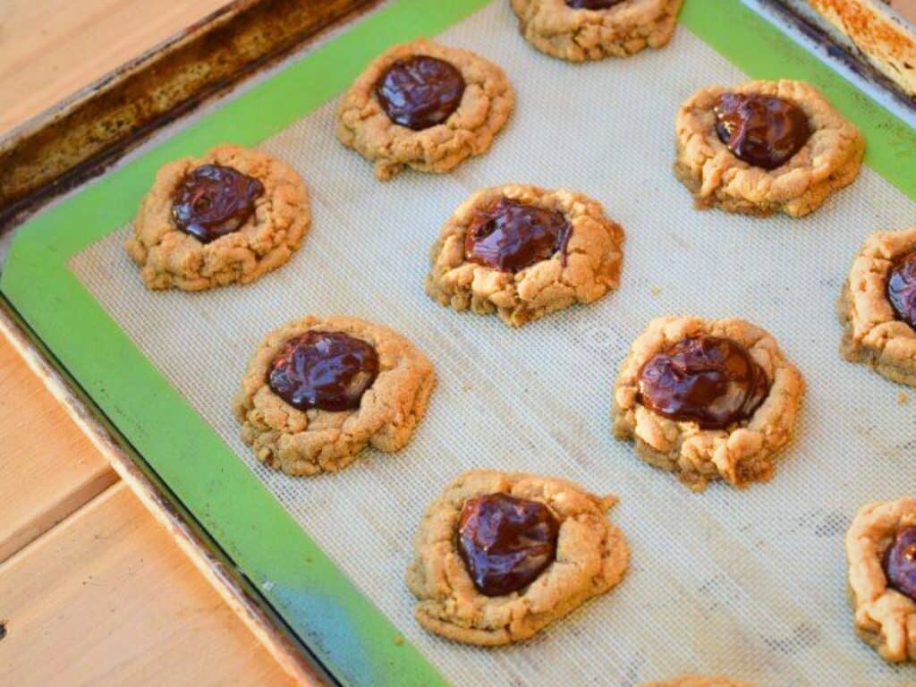 A baking tray with nine s&rsquo;mores cookies, each peanut butter cookie topped with a dollop of melted chocolate, on a silicone mat.