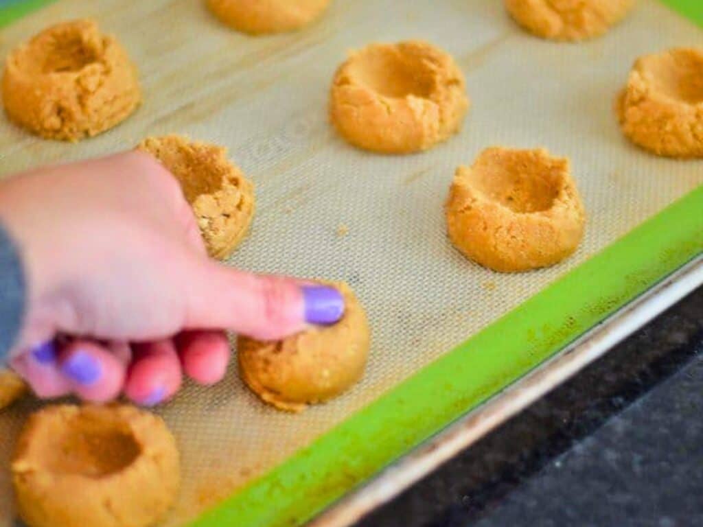 A hand presses a thumbprint into unbaked s&rsquo;mores cookies dough balls on a baking sheet lined with a silicone mat.