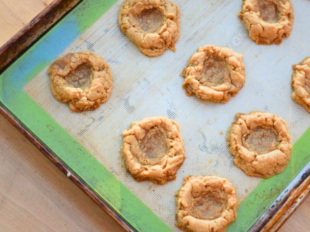 A baking sheet with seven s&rsquo;mores cookies, each with a deep indent in the center, sits on a wooden surface.