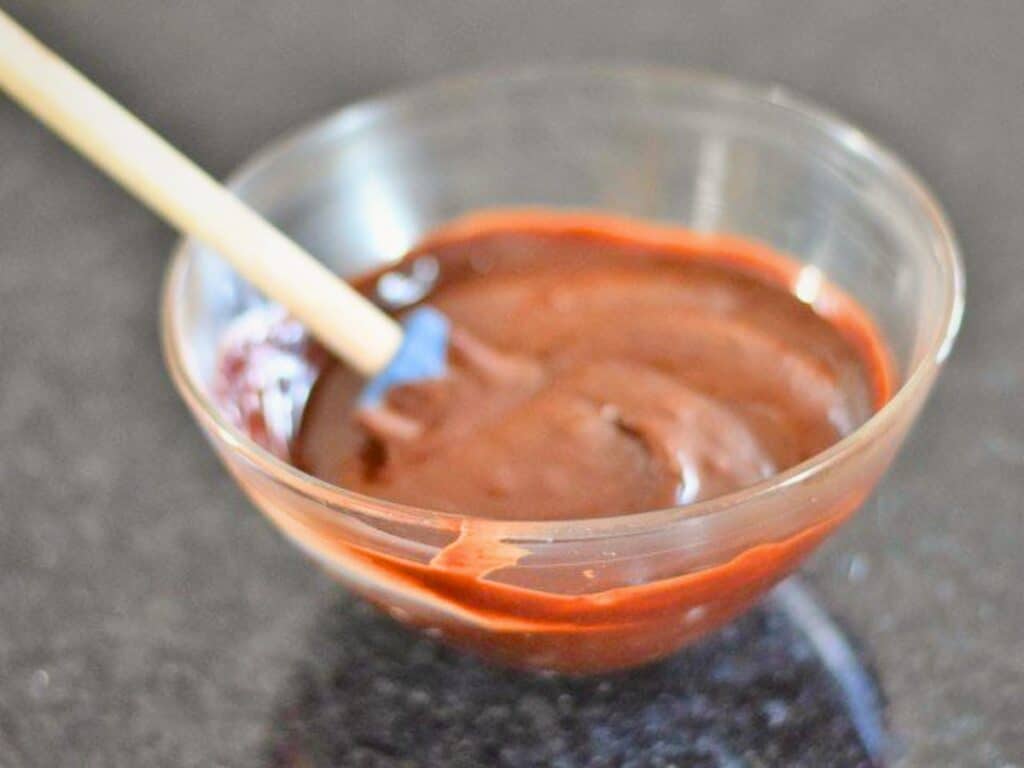 A clear glass bowl containing chocolate batter for s&rsquo;mores cookies, with a spatula resting inside, placed on a dark countertop.