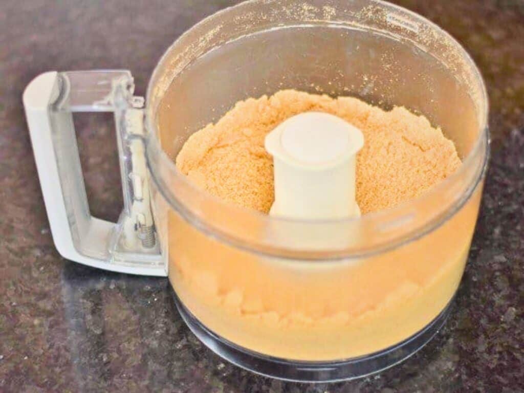 A food processor bowl containing finely ground orange-brown crumbs for s&rsquo;mores cookies sits on a dark countertop.