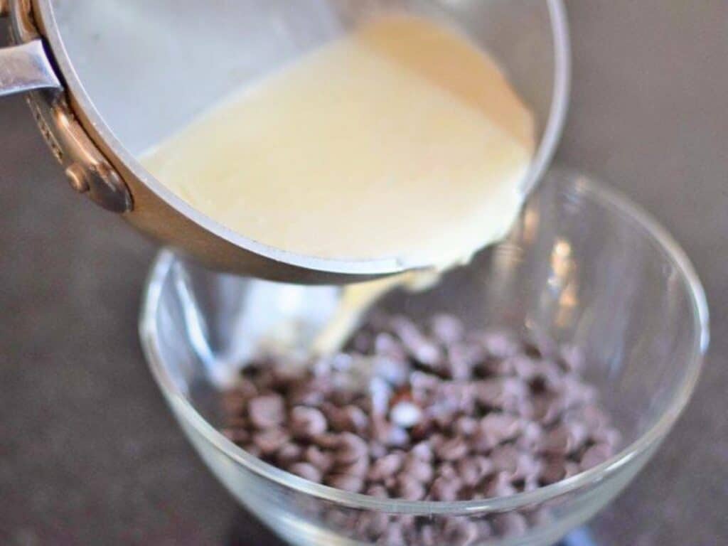 A pot of cream is being poured over a bowl of chocolate chips, likely to make ganache for s&rsquo;mores cookies, on a kitchen countertop.