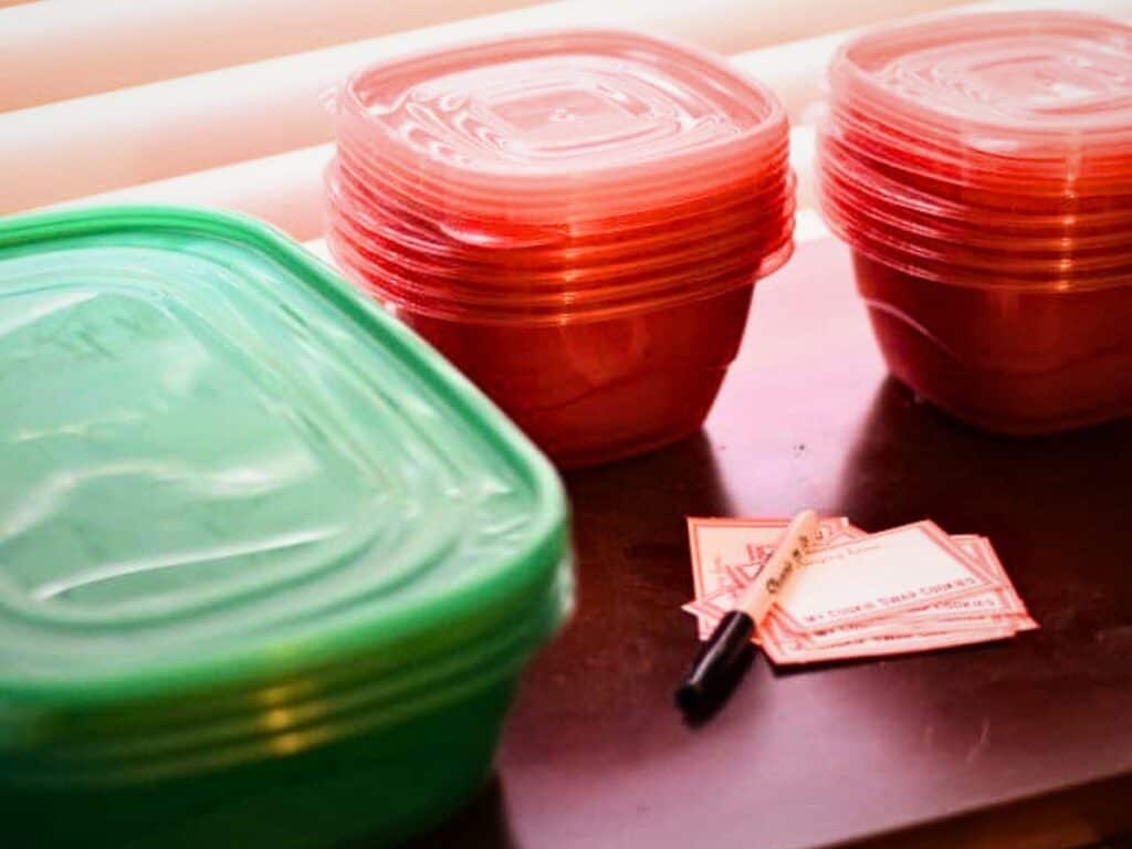 Plastic food storage containers stacked on a table next to a pen and blank food labels, perfect for organizing fresh batches of s&rsquo;mores cookies.