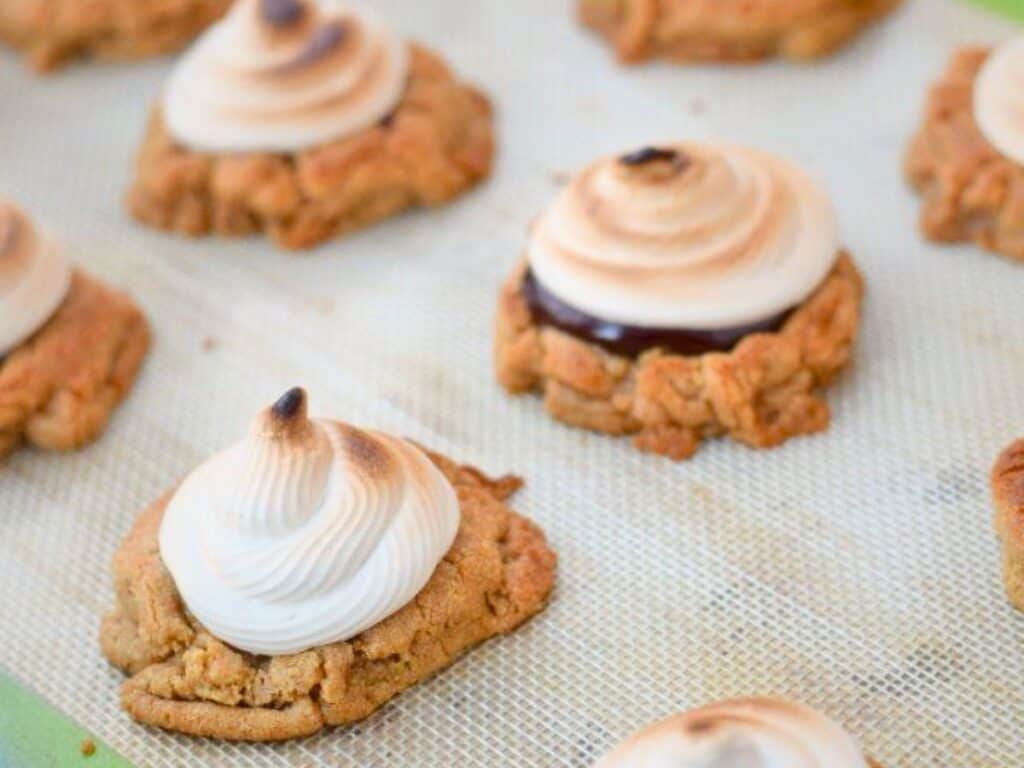 Peanut butter s&rsquo;mores cookies topped with swirled, toasted meringue sit on a silicone baking mat, some with a layer of chocolate beneath the meringue.