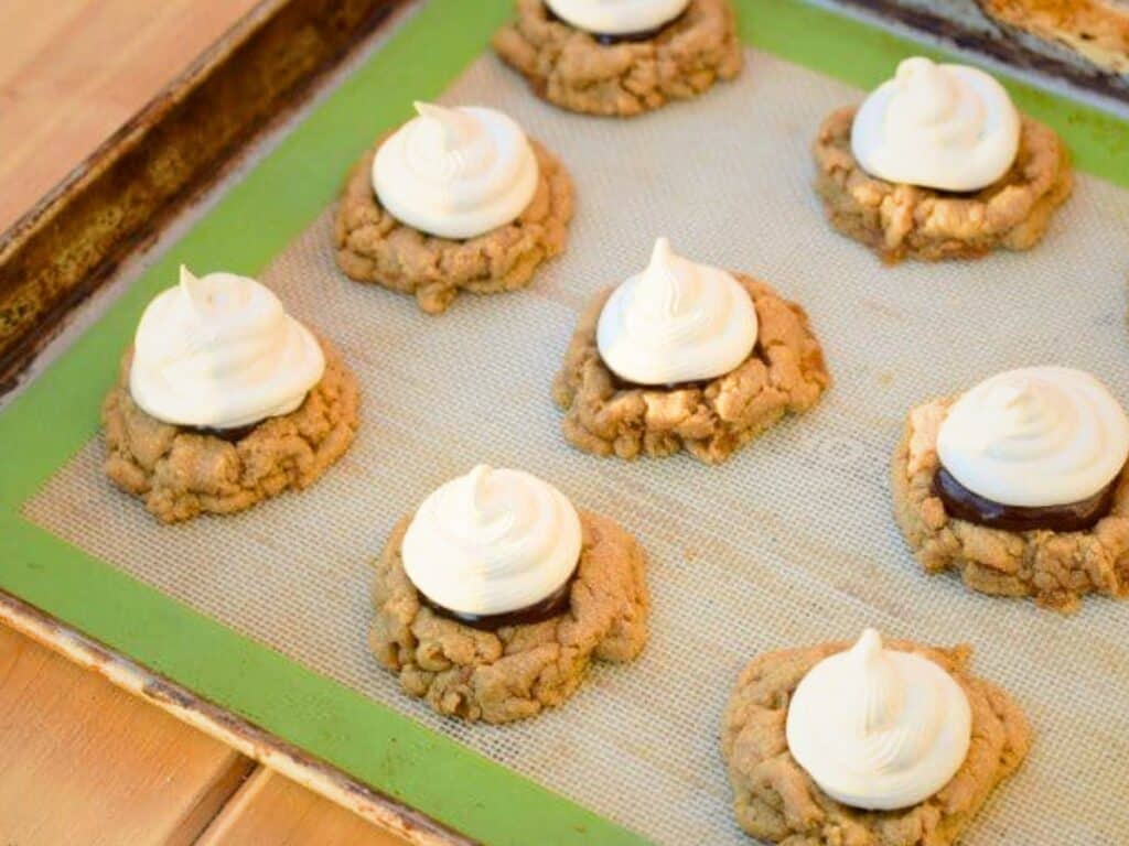 A baking sheet with eight s&rsquo;mores cookies topped with white frosting, sitting on a silicone baking mat.