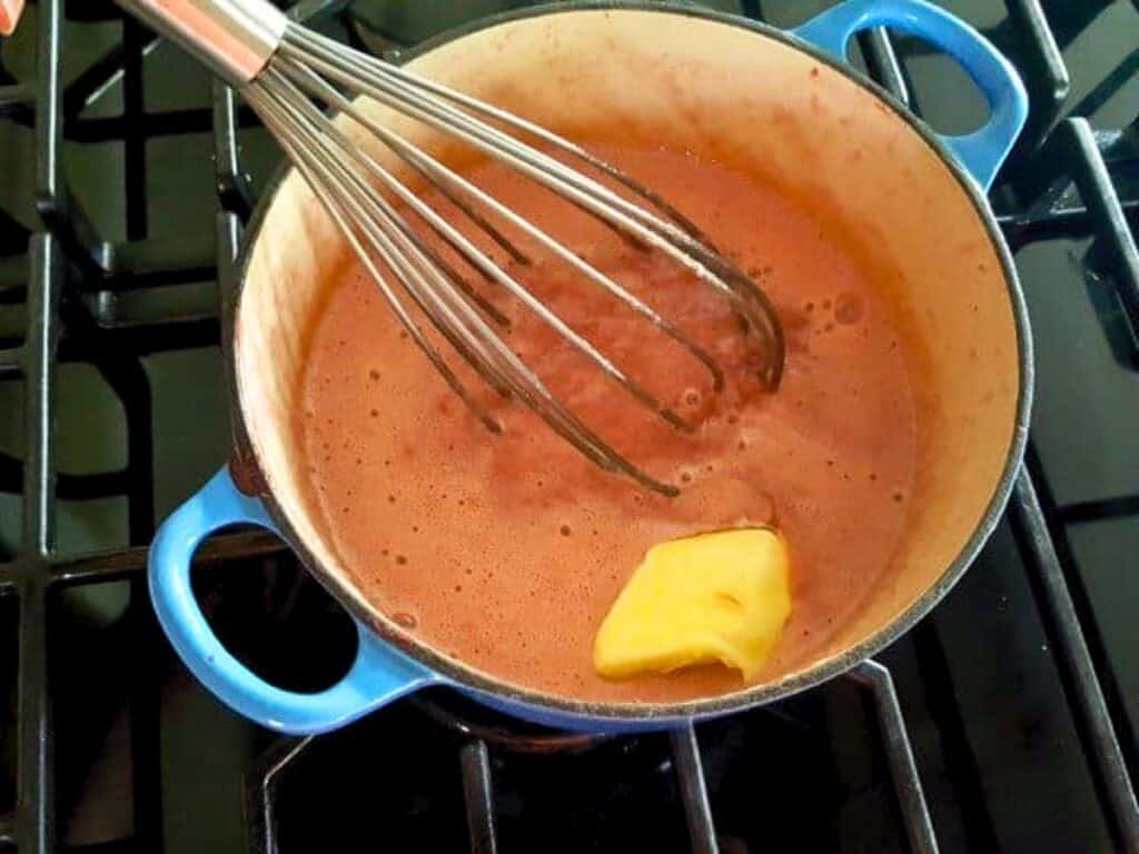 A blue pot on a stove contains a chocolate mixture being whisked, with a piece of butter melting in the mixture, ready to be layered with homemade strawberry curd for a decadent dessert.
