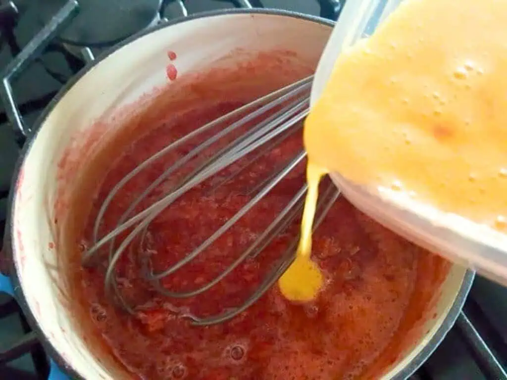 Egg mixture is being poured into a saucepan of red liquid on a stove, with a whisk mixing the contents to create a smooth strawberry curd.