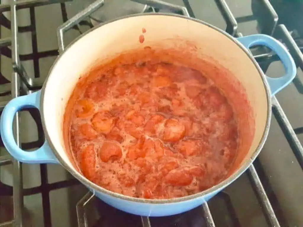 A blue pot on a stovetop contains a simmering, chunky red fruit mixture, likely strawberry jam or strawberry curd, being cooked.