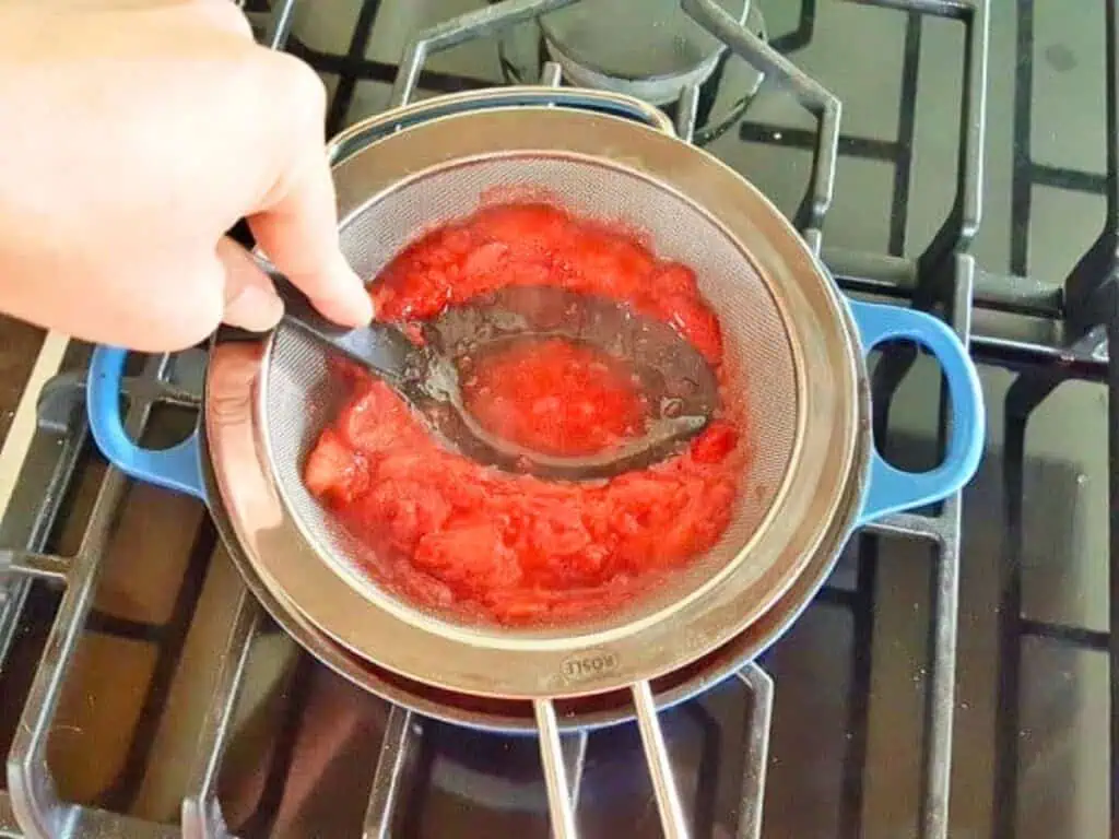 Hand pressing stewed tomatoes through a metal strainer with a black spoon over a blue pot on a gas stove, much like the method used when preparing silky homemade strawberry curd.