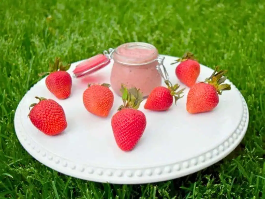 A white plate on grass holds several strawberries arranged around a small jar of strawberry curd dip with a pink spoon resting on top.