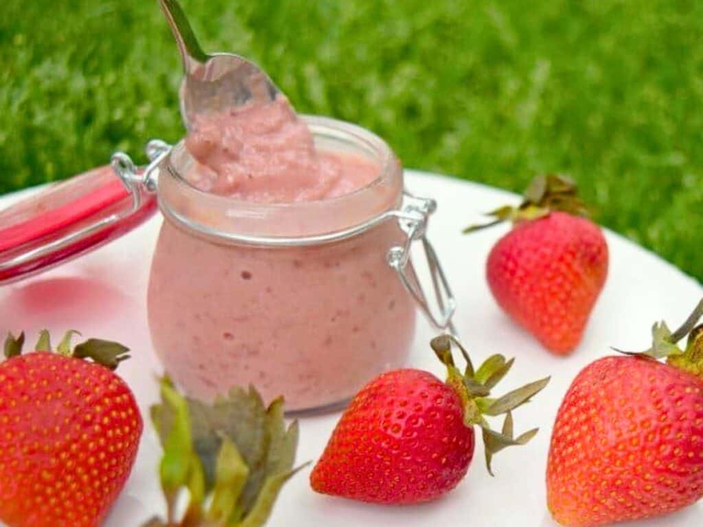A small glass jar of strawberry mousse with a spoon inside is placed on a white plate surrounded by fresh strawberries on a grassy background, featuring luscious layers of strawberry curd for an extra burst of flavor.