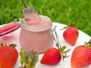 A small glass jar of strawberry mousse with a spoon inside is placed on a white plate surrounded by fresh strawberries on a grassy background, featuring luscious layers of strawberry curd for an extra burst of flavor.
