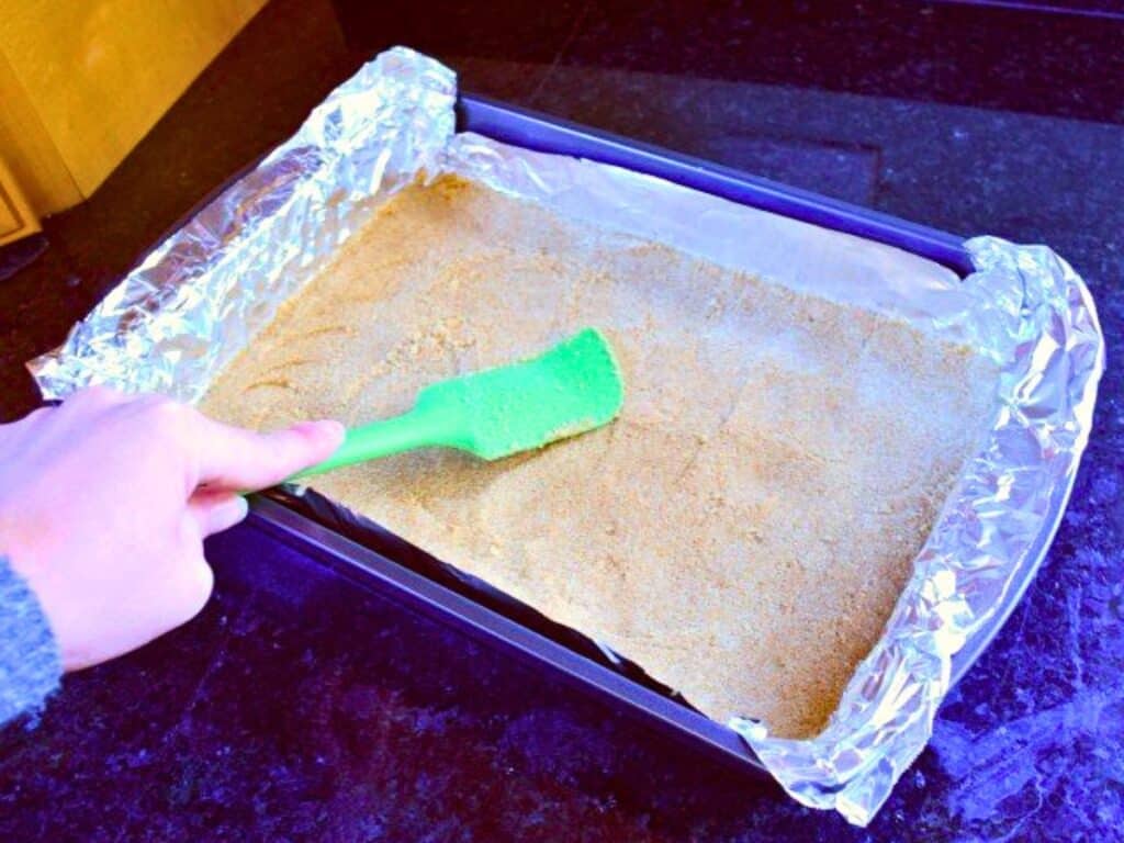 A hand uses a green spatula to press graham cracker crust for s&rsquo;mores brownies into a foil-lined rectangular baking pan on a dark countertop.