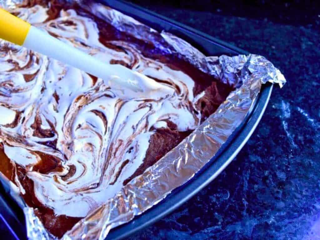 A baking pan lined with foil contains marbled s&rsquo;mores brownie batter being swirled with a spatula on a dark countertop.