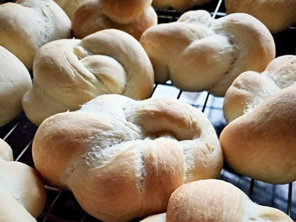 Close-up of several homemade dinner rolls twisted into elegant shapes, cooling on a wire rack. The rolls have a light golden crust and a soft, fluffy texture.