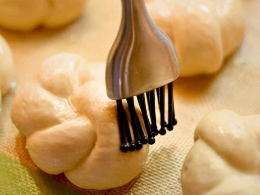 A silicone brush is applying an egg wash to unbaked, braided homemade dinner rolls on a baking sheet.