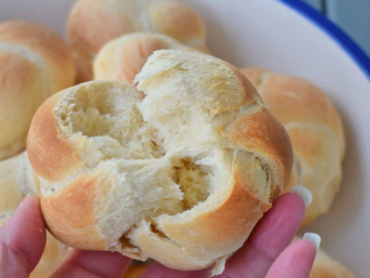 A hand holds a freshly baked, partially torn homemade dinner roll with a soft interior. More similar rolls are visible in a bowl in the background.