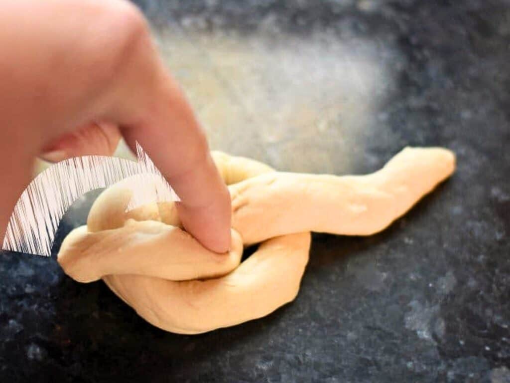 A hand shapes dough into a knot on a dark countertop, preparing homemade dinner rolls, with an arrow indicating the direction to tuck the dough end.