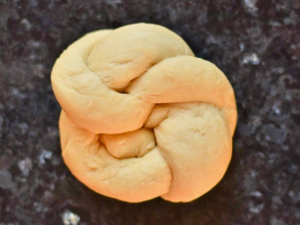A single piece of braided, unbaked bread dough is shaped into a knot and placed on a dark, speckled countertop, ready to be baked into delicious homemade dinner rolls.