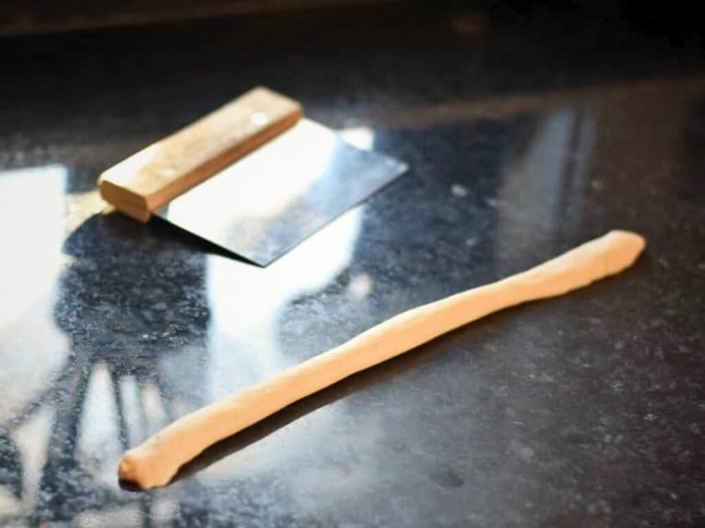 A metal bench scraper and a rolled log of dough for homemade dinner rolls rest on a dark countertop.