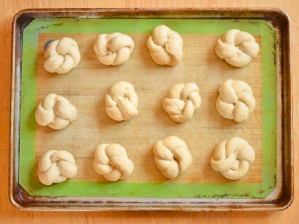 Twelve unbaked knotted dough pieces, perfect for homemade dinner rolls, are arranged evenly on a baking sheet lined with a silicone mat.