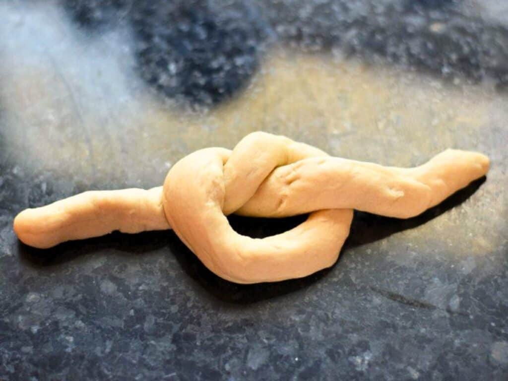 A piece of dough shaped into a knot, soon to become delicious homemade dinner rolls, rests on a dark countertop.