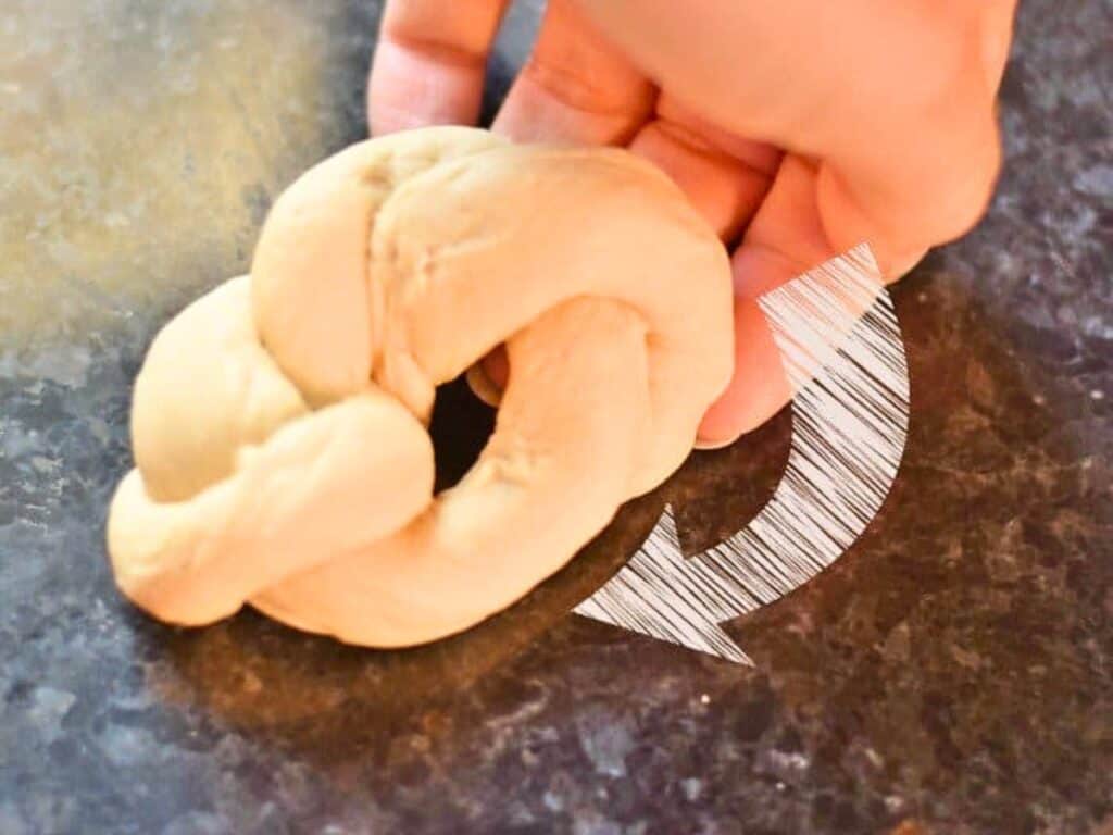 A hand twists a piece of dough into a knot shape on a dark countertop, preparing homemade dinner rolls, with a white arrow graphic indicating the twisting motion.