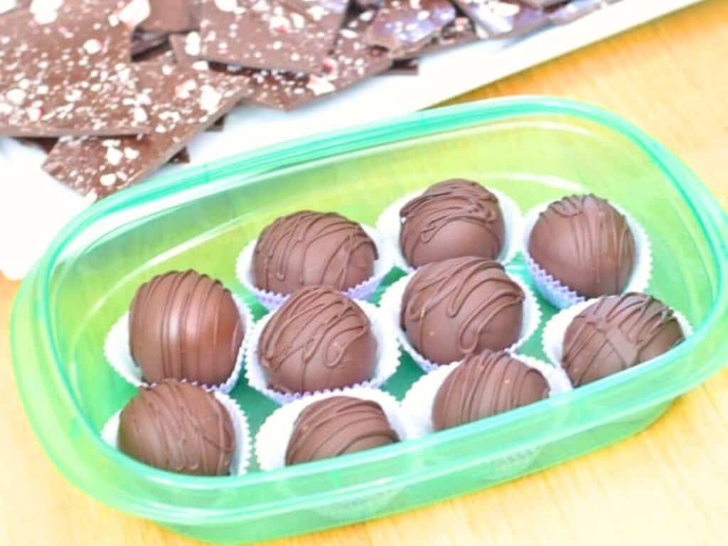 Nine chocolate truffles with drizzled chocolate on top, arranged in paper cups inside a green plastic container. Brown sugar cookie dough truffles nestle alongside classic flavors, while brown chocolate bark pieces are visible in the background.