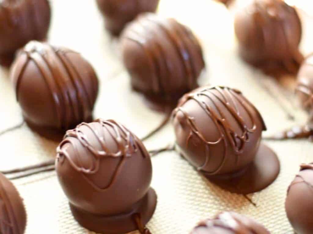 Rows of round brown sugar cookie dough truffles covered in smooth chocolate coating and drizzled with more chocolate, placed on a textured surface.