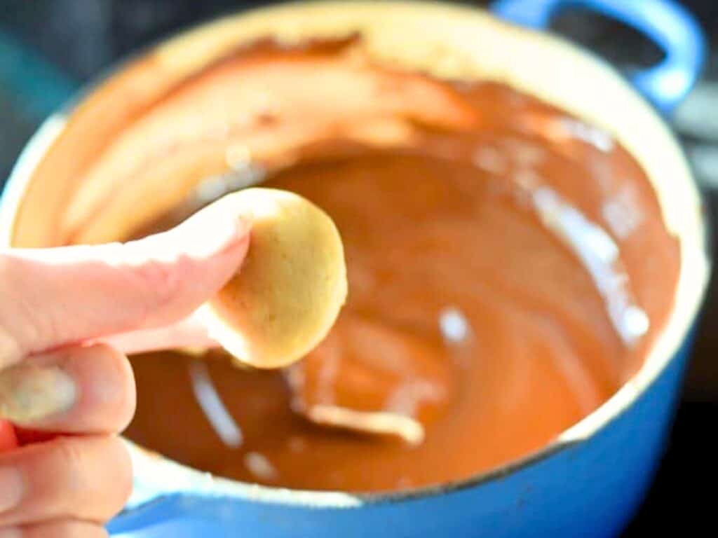 A hand holding a small round dough ball over a blue pot filled with melted chocolate, preparing to create delectable brown sugar cookie dough truffles.