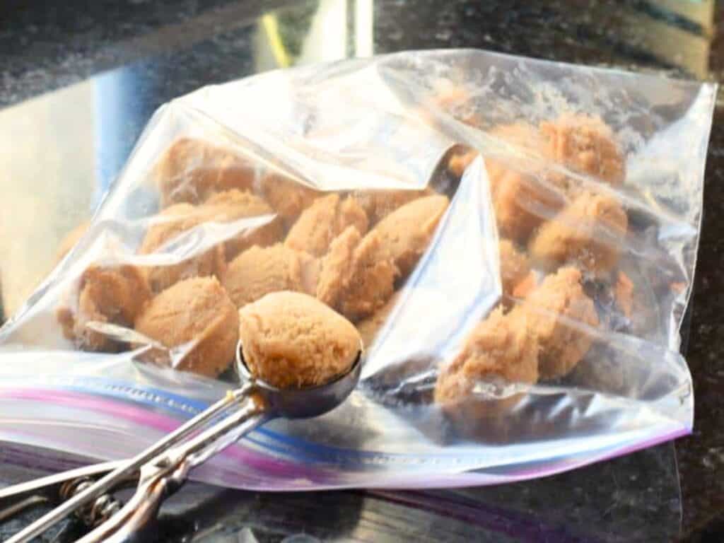 A resealable plastic bag filled with scooped balls of brown sugar cookie dough truffles sits on a dark countertop, with a metal cookie scoop resting nearby.