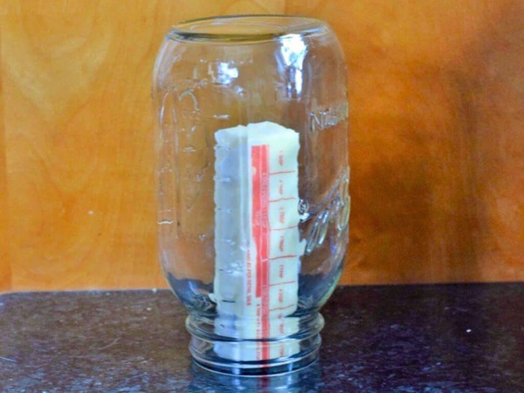 An upside-down glass jar covers a stick of butter standing upright on a dark countertop with a wooden background, reminiscent of preparing brown sugar cookie dough truffles in a cozy kitchen setting.