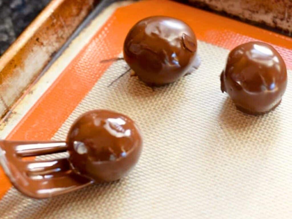 Three round brown sugar cookie dough truffles sit on a silicone baking mat, with one being coated in chocolate using a dipping tool.