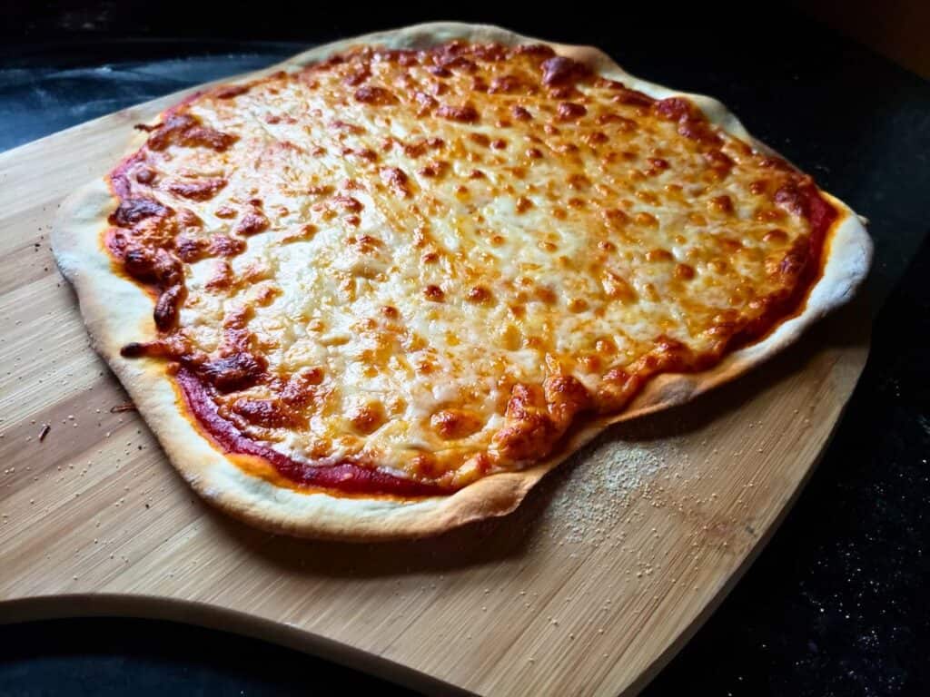 A homemade cheese pizza with a thin, irregular crust made from homemade pizza dough and melted cheese sits on a wooden pizza peel.