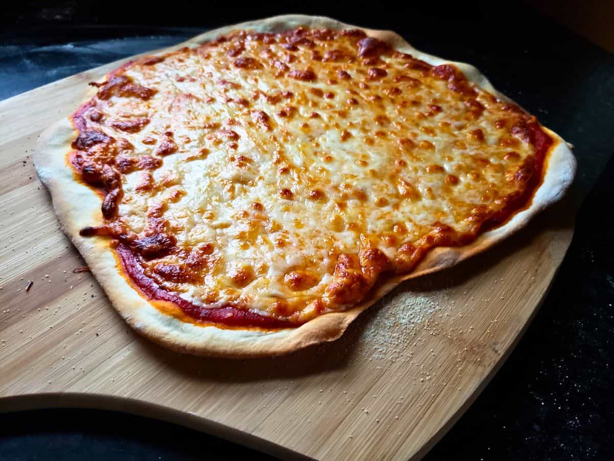 A homemade cheese pizza with a thin, irregular crust made from homemade pizza dough and melted cheese sits on a wooden pizza peel.