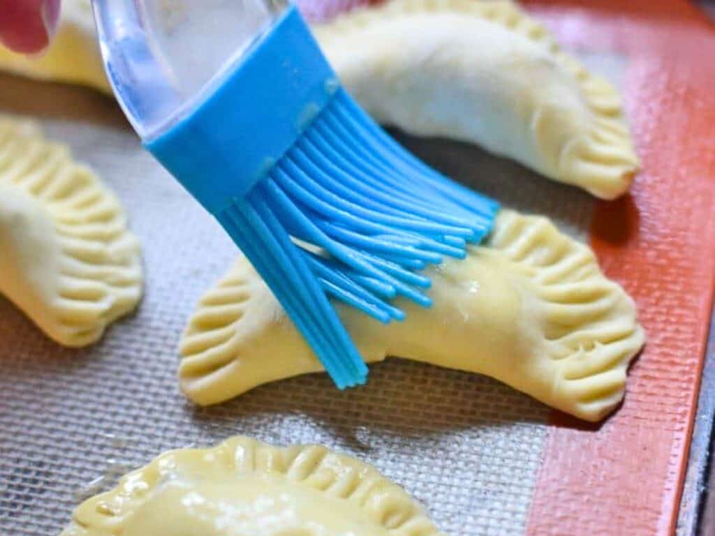 A blue silicone brush applies an egg wash to uncooked, crimped pastry turnovers on a baking sheet.
