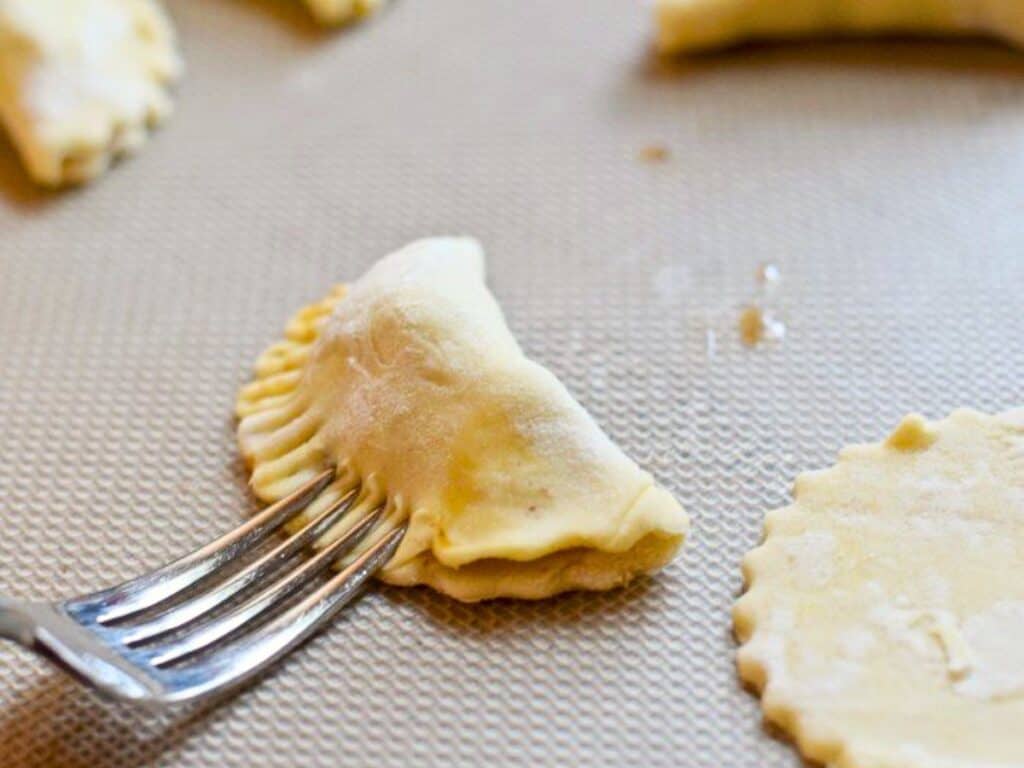 A fork is pressing the edges of a semicircular dough pocket to seal it; uncooked dough rounds are nearby on a baking sheet.