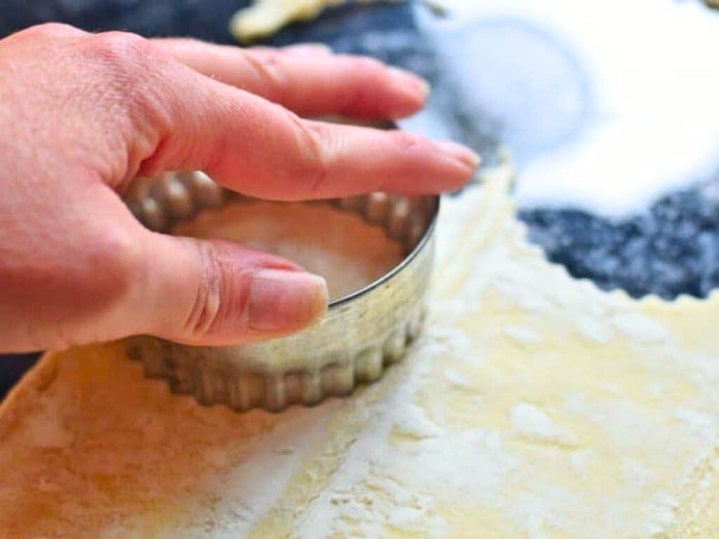 A hand presses a round, fluted biscuit cutter into rolled-out dough on a floured surface.