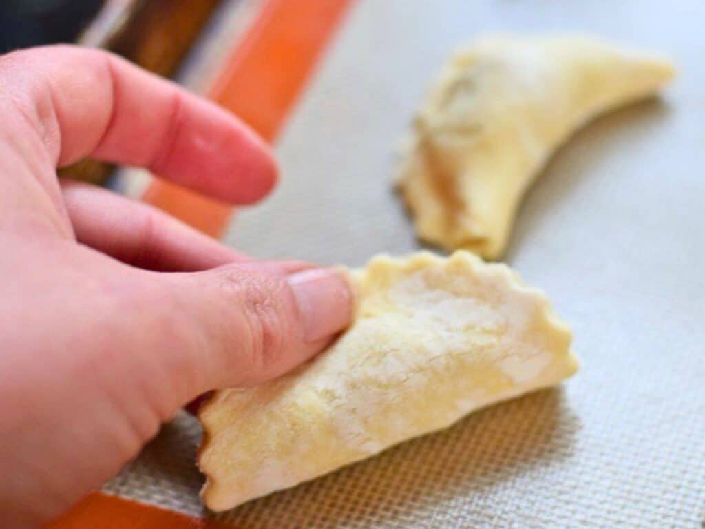 A hand holds an uncooked, crimped pastry over a baking mat, with another similar pastry resting in the background.