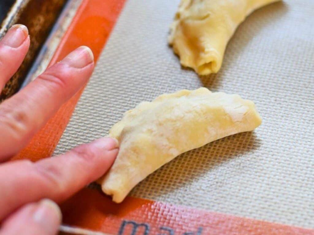 A hand presses the edge of an uncooked, folded pastry on a silicone baking mat, with another similar pastry in the background.
