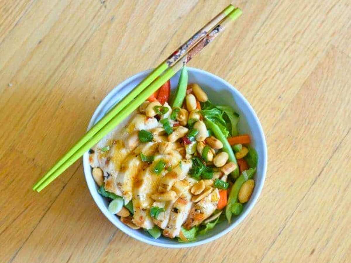 A vibrant bowl of kung pao chicken salad featuring grilled chicken, peanuts, crisp vegetables, and green onions, all lovingly topped with a savory sauce. A pair of green chopsticks rests on the bowl.