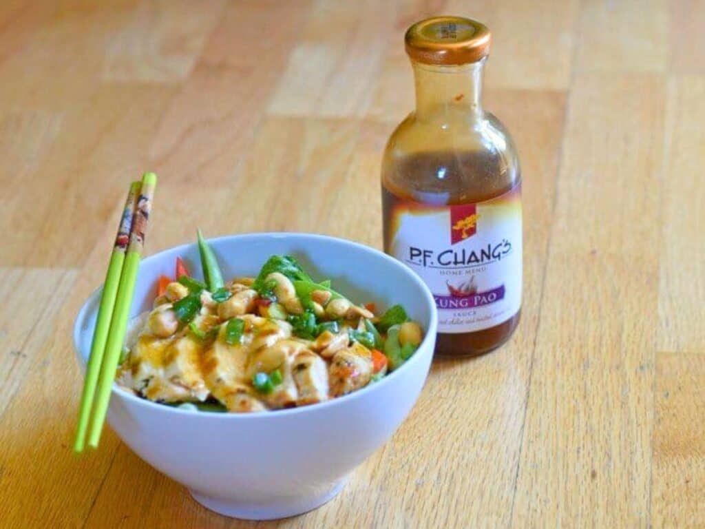 A bowl of stir-fry with vegetables and sauce sits next to a bottle of P.F. Chang&rsquo;s Kung Pao sauce on a wooden surface, with green chopsticks resting on the bowl&mdash;a fresh twist for creating your own kung pao chicken salad at home.