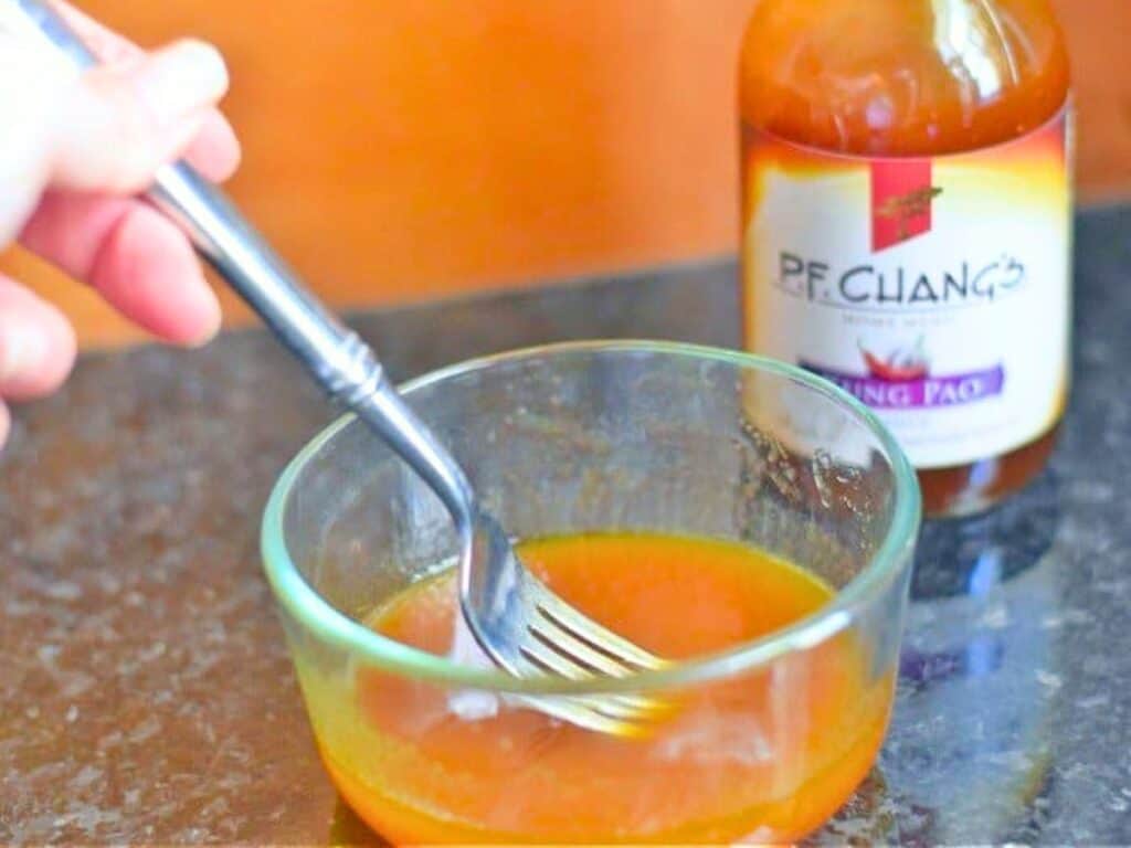 A hand mixes orange sauce in a glass bowl with a fork; a bottle of P.F. Chang's Kung Pao sauce is next to the bowl, perfect for preparing a flavorful kung pao chicken salad.