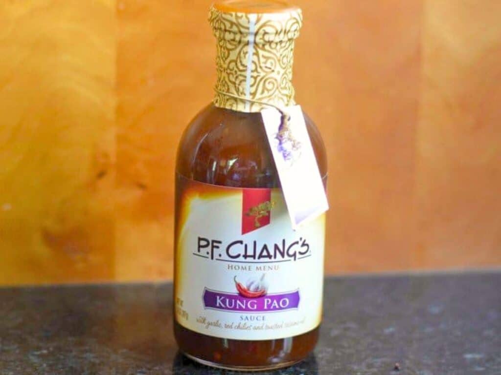 A bottle of P.F. Chang's Kung Pao sauce sits on a dark countertop against a wood background, with a gold patterned cap and a hanging tag&mdash;perfect for adding bold flavor to your next kung pao chicken salad.
