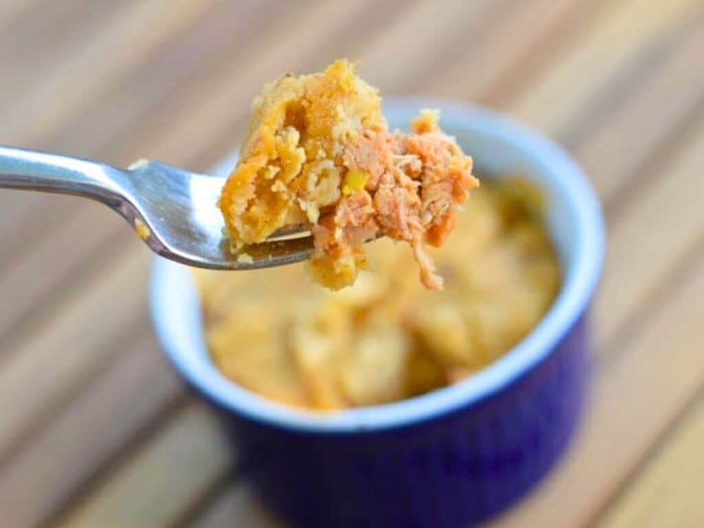 A fork holds a bite of a baked dish with shredded meat and crust, with a small blue ramekin of tamale pot pies in the background.